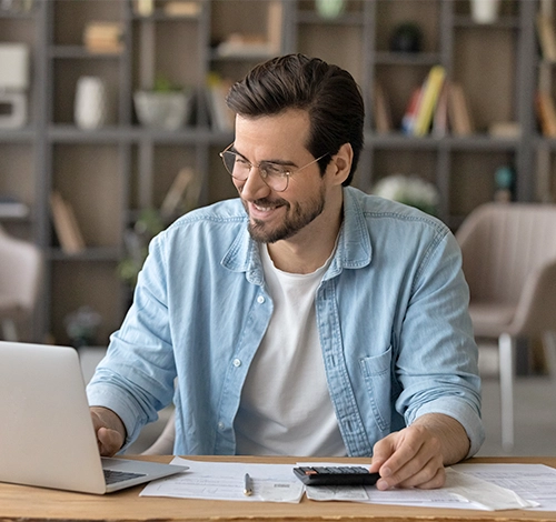BCA-Leistungen Ein Mann sitzt im Büro vor Laptop mit Papierunterlagen und Taschenrechner und blickt zufrieden auf den Monitor.