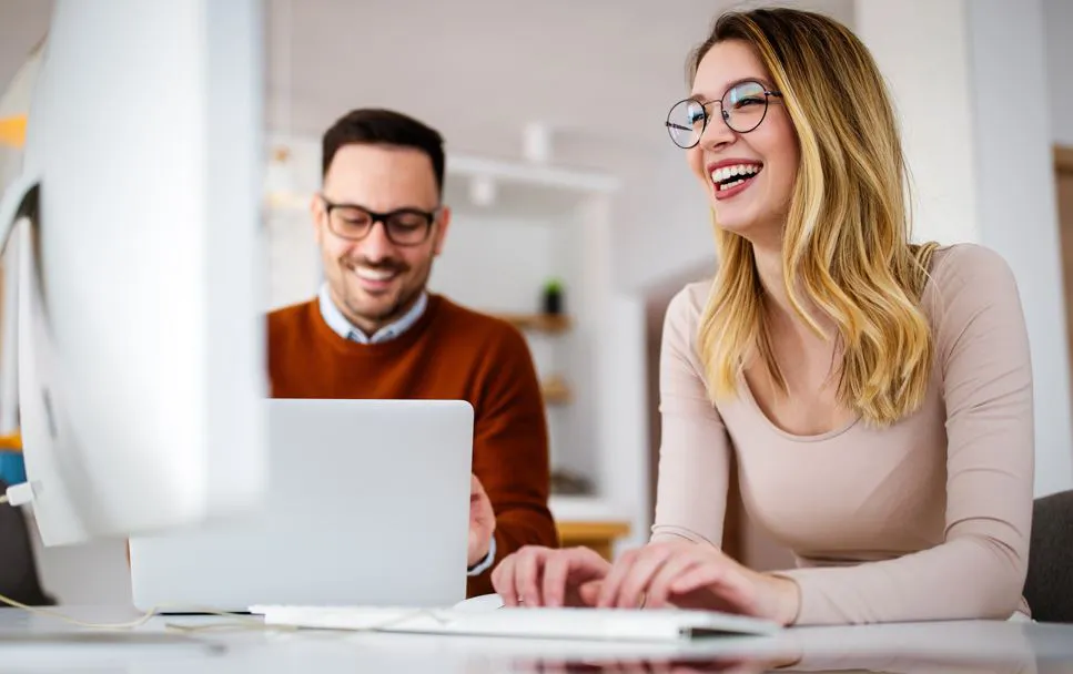 Eine föhliche Mitarbeiterin und ihr Kollege arbeiten vor dem Computer in einem hellen Büro