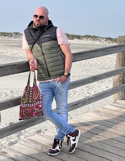 Ein Mann mit kurzärmeligem Hemd, Weste und Jeans hält eine farbenfrohe Tasche.