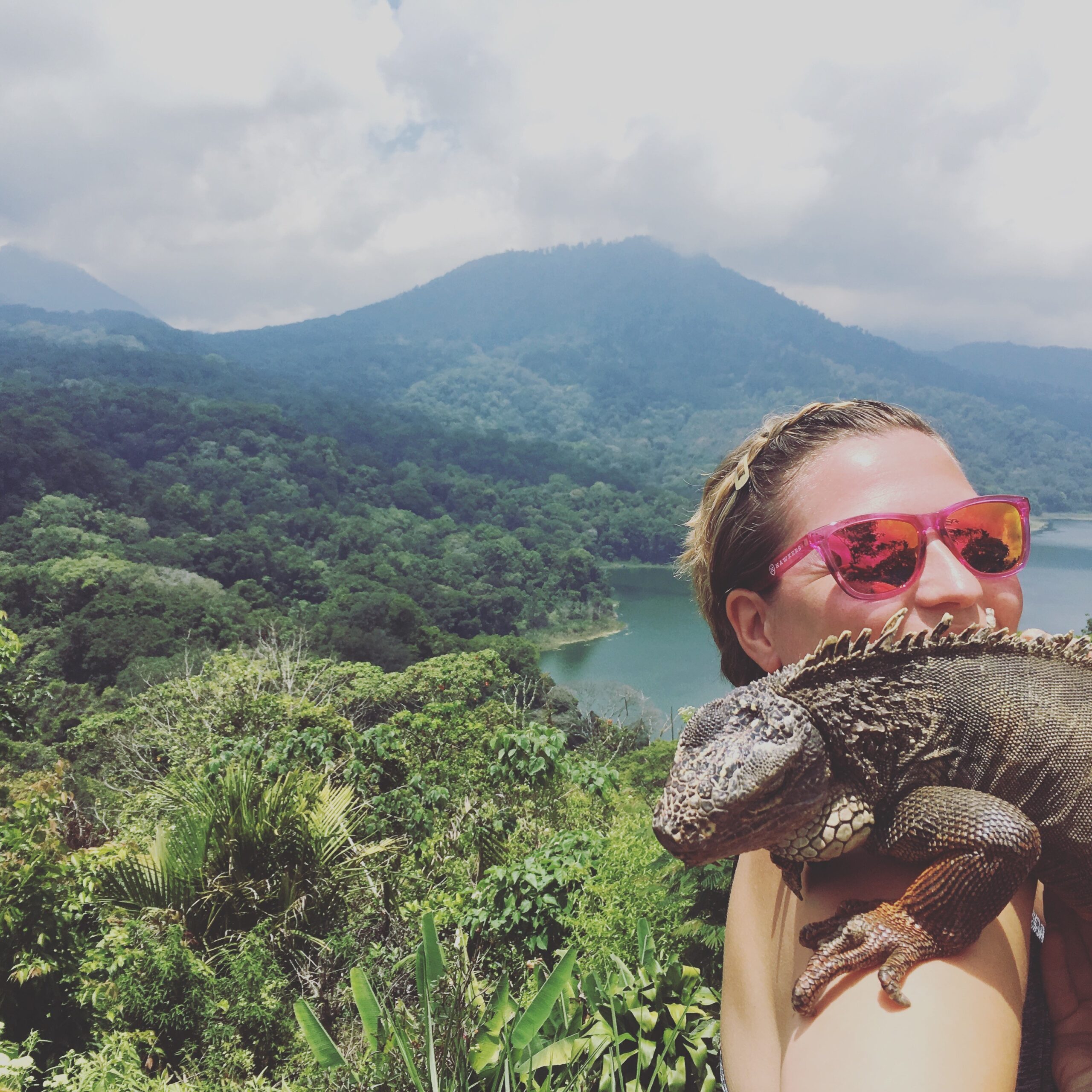 Ein Mädchen hält einen großen Leguan in den Armen. Im Hintergrund eine wunderschöne Naturkulisse.