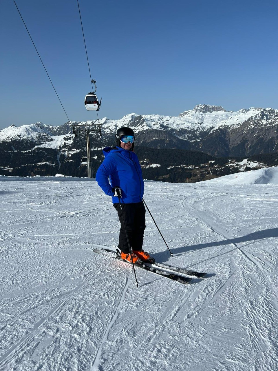 Ein Skifahrer steht auf der Piste und blickt zur Bergbahn. Er trägt eine blaue Jacke und schwarze Hose.