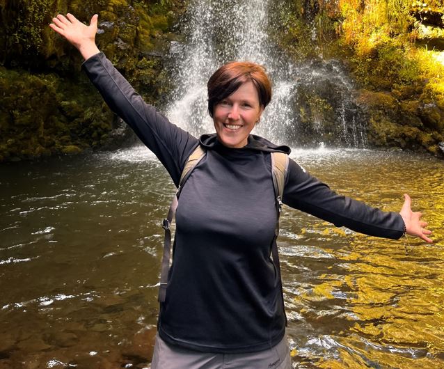 Eine Frau steht vor einem Wasserfall. Sie hat ihre Arme ausgebreitet.
