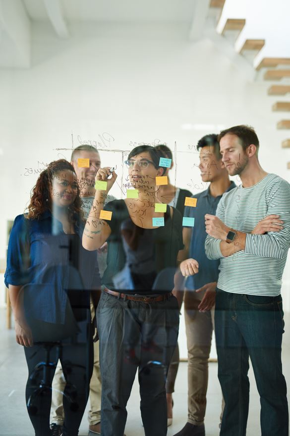 Gruppe von Kollegen beim Brainstorming mit Post-it-Notizen Ein Team von Erwachsenen steht zusammen und betrachtet bunte Klebenotizen an einer Glasscheibe. Sie scheinen im Rahmen eines Meetings Ideen zu diskutieren.