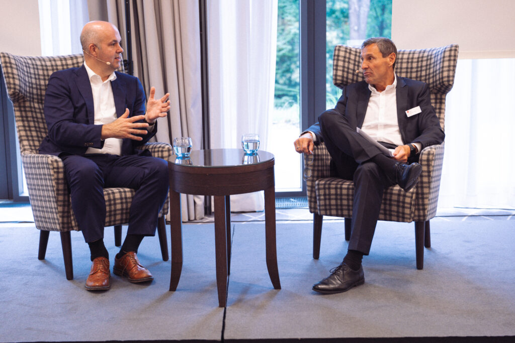 Zwei Geschäftsleute unterhalten sich in einem Raum. Sie sitzen auf großen, gemütlichen Stühlen.