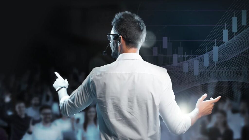 Ein Mann in einem weißen Hemd zeigt auf eine Grafik. Er steht vor einem Publikum.