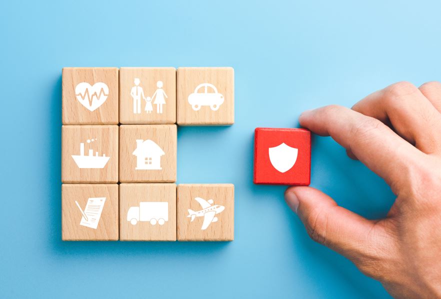 Ein Hand hält ein rotes Holzstück mit einem Schildsymbol. Holzstücke mit Versicherungssymbolen liegen daneben.