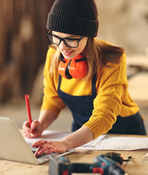 Eine Frau in gelber Bluse und schwarzer Mütze arbeitet am Laptop. Sie trägt Ohrenschützer und schreibt Notizen.