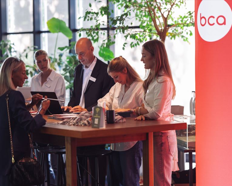 Ein Team von fünf Personen steht um einen Tisch herum, ein großes Fenster mit Bäumen im Hintergrund. Das bca Logo ist sichtbar.