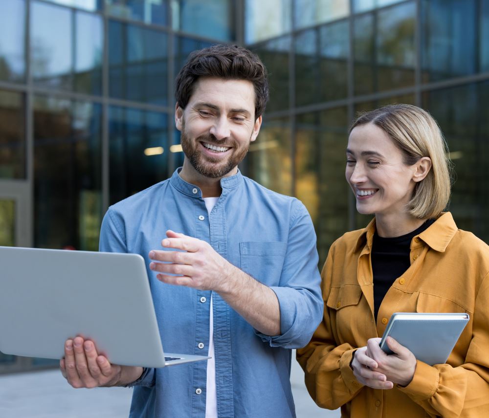 Ein Mann und eine Frau stehen vor einem großen Fenster und betrachten einen Laptop und ein Tablet. Sie scheinen zu diskutieren.