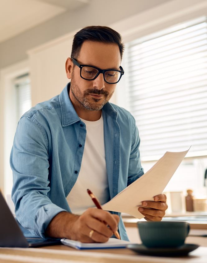 Ein Mann mit Brille und blauem Hemd schreibt auf einem Papier. Er sitzt an einem Tisch.