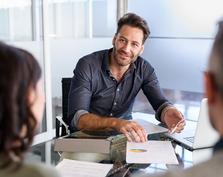 Ein Mann sitzt an einem Tisch und präsentiert Unterlagen. Er lächelt und ist in einer Besprechung.