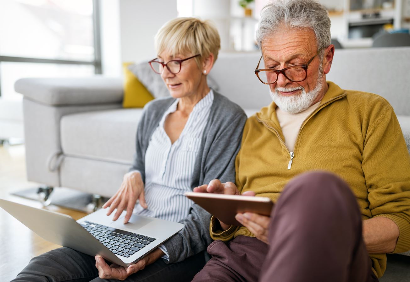 Ein älteres Paar sitzt auf einem Sofa und nutzt einen Laptop und ein Tablet.