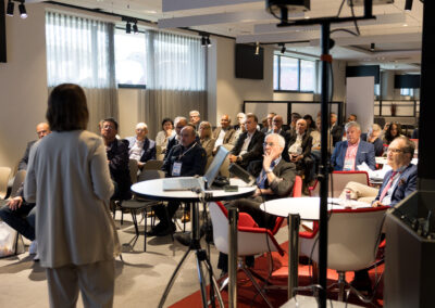 Eine Frau steht vor einem Publikum in einem Konferenzraum. Die Zuhörer sitzen an runden Tischen.