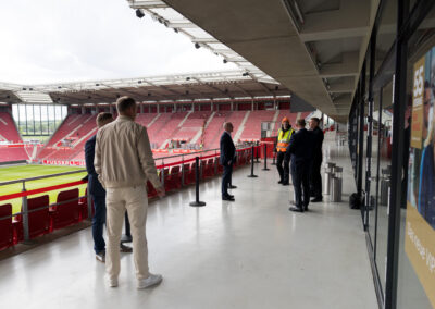 Einige Männer stehen auf den Gängen eines leeren Fußballstadions mit roten Sitzen.