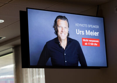 Ein Mann in einem dunklen Hemd auf einem Bildschirm. Der Text daneben nennt ihn als Keynote-Speaker.