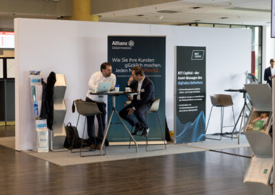 Ein Messe-Stand mit zwei Männern, die an einem Tisch sitzen. Der Stand hat eine weiße Wand und zwei blaue Werbeflächen.