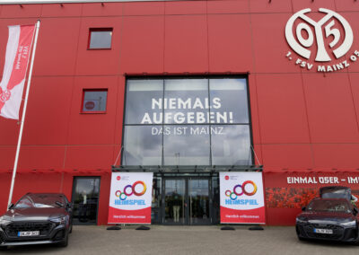 Ein rotes Gebäude mit zwei Autos davor und einer Flagge links. Das Gebäude hat eine große Scheibe mit Text.