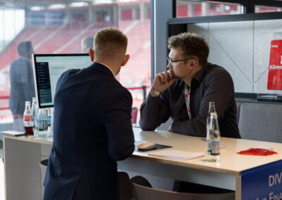 Zwei Männer sitzen an einem Tisch und unterhalten sich. Im Hintergrund sind rote Sitzreihen sichtbar.