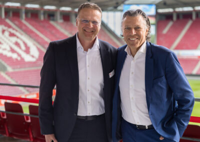 Zwei Männer lächelnd im Stadion. Sie tragen Anzüge.