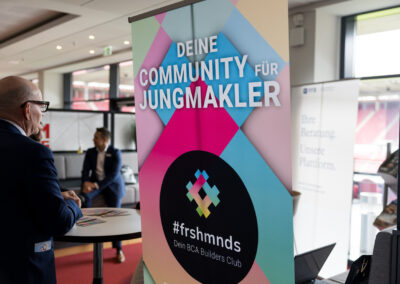 Ein Mann steht vor einem Tisch, im Hintergrund eine Frau. Ein großes Logo mit dem Slogan 'Deine Community für Jungmakler' ist sichtbar.