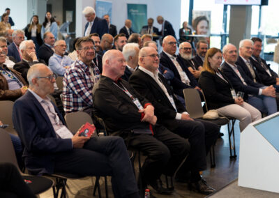Eine Gruppe von älteren Männern in formeller Kleidung sitzt in einem Konferenzraum. Sie scheinen auf einen Vortrag zu warten.