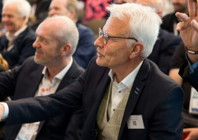 Ein älterer Mann mit weißem Haar und Brille in einem dunklen Anzug. Ein anderer Mann im Hintergrund.