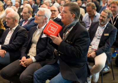 Ein Mann in einem dunklen Jackett hält ein rotes Buch. Er sitzt unter anderen Zuhörern in einem Saal.