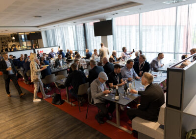 Ein Meeting mit vielen Teilnehmern in einem hellen Raum. Die Leute sitzen an Tischen und hören zu.