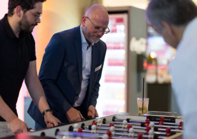 Ein Mann im schwarzen Hemd und ein weiterer im blauen Anzug spielen gegen einen Mann im weißen Hemd. Der Spieler im Anzug blickt konzentriert auf das Spiel.