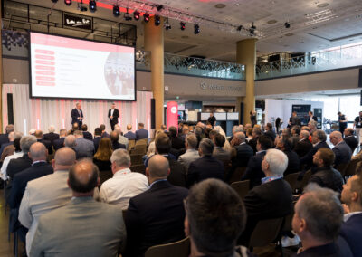 Ein großer Saal voller Männer in Anzügen, die einer Präsentation zuhören. Die Bühne hat eine große Leinwand.