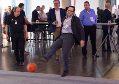 Ein Mann in einem Anzug kickt einen orangefarbenen Ball. Andere Männer im Hintergrund beobachten ihn.
