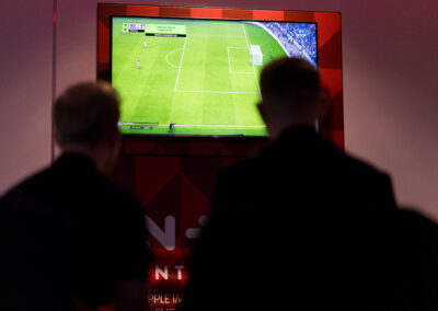 Zwei Männer beobachten ein Fußballspiel auf einem Bildschirm. Sie stehen vor einem großen, roten Rahmen.