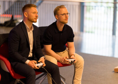 Zwei Männer sitzen auf roten Stühlen und spielen ein Videospiel. Sie sind beide auf die Bildschirme vor ihnen konzentriert.