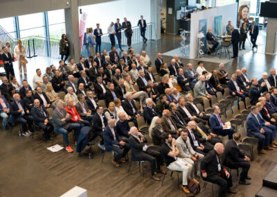 Ein Saal voller Menschen in Businesskleidung. Sie sitzen auf Stühlen und hören einem Vortrag zu.