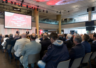 Ein Auditorium mit Männern, die Vortrag hören. Sie sitzen auf Stühlen und schauen auf eine große Leinwand.