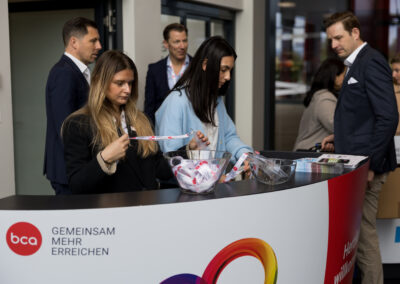 Ein paar junge Erwachsene stehen an einem Tisch, während eine Frau einen Gegenstand in der Hand hält. Einige andere Personen im Hintergrund unterhalten sich.