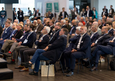 Ein Saal voller Männer in Businesskleidung. Sie sitzen auf Stühlen und hören einem Vortrag zu.
