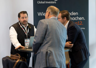 Drei Männer in Anzügen sprechen miteinander auf einer Messe. Ein Mann sitzt und erklärt etwas.