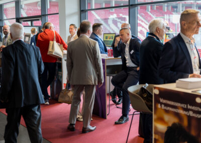 Männer in Anzügen unterhalten sich auf einer Messe. Sie stehen um Tische herum.